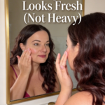 Close-up of a woman at a bathroom mirror touching her cheek while the image text says makeup that looks fresh not heavy.
