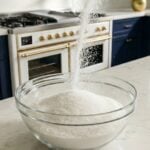 Sugar being poured into a bowl on a white quartz countertop in a dark blue and gold kitchen.