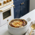 Steaming bowl of onion boil with bone broth topped with melted cheese in a white ramekin on a white quartz countertop with dark blue cabinets and gold hardware in the background.
