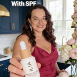 Woman smiling in a modern kitchen with dark blue cabinets holding a skincare-makeup bottle forward, text reads over-50 glow starts with SPF.