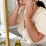 Woman washing her face at a bathroom vanity with cleanser foam on her hands, product bottle on the counter.