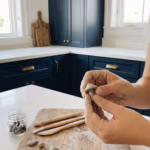 Hands sculpting a small clay mushroom magnet on a craft table with clay tools, cutters, and unfinished clay shapes in the background.