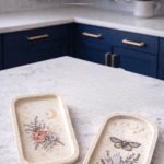 Two decorative clay trays with floral and botanical transfer art styled on a white quartz countertop in a bright blue kitchen.
