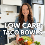Low carb high protein taco bowl held by a smiling woman in a blue kitchen, topped with ground beef, shredded cheese, lettuce, salsa, avocado, and sour cream.