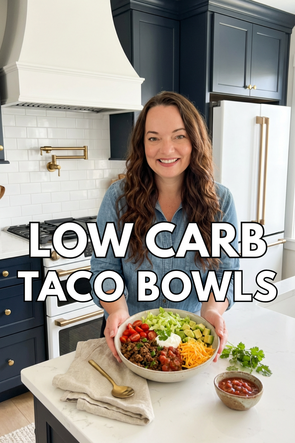 Low carb high protein taco bowl held by a smiling woman in a blue kitchen, topped with ground beef, shredded cheese, lettuce, salsa, avocado, and sour cream.
