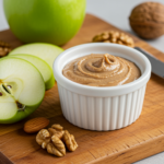 Green apple slices with a white ramekin of nut butter on a wooden cutting board, surrounded by walnuts and almonds.