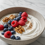Bowl of plain Greek yogurt topped with raspberries, blueberries, walnuts, chia seeds, and cinnamon on a marble countertop.