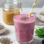 Pink berry smoothie in a glass topped with chia seeds and pumpkin seeds, surrounded by spinach, almonds, and frozen berries.
