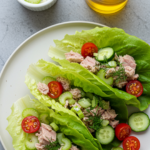 Romaine lettuce wraps filled with tuna salad, celery, cucumber slices, dill, and cherry tomatoes on a white plate.