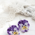 Handmade clay pansy earrings in purple, white, and yellow displayed on a white rustic surface with lace, styled as floral spring jewelry.