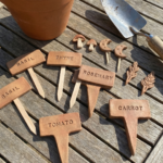 Handmade clay garden markers labeled basil, thyme, rosemary, tomato, and carrot arranged on a wooden table beside a herb pot and gardening trowel.