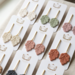 Multiple pairs of polymer clay leaf earrings in neutral and pastel colors displayed on branded jewelry cards near a window.