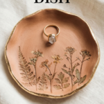 Round terracotta clay ring dish with pressed flower designs and a gold rim, holding a ring on soft neutral linen.