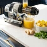 Nama cold pressed juicer sitting on a white quartz counter with dark blue cupboards with gold hardware in the background. A mix of fruit sits on the counter beside it.