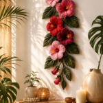 Crocheted hibiscus tapestry wall hanging with large red and pink yarn flowers and green leaves mounted on a white wall above a rattan shelf with tropical plants and candles in warm golden afternoon light