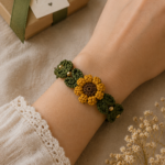 Close-up of a crocheted sunflower bracelet in yellow and brown yarn worn on a wrist resting on beige linen beside a kraft paper gift box with a green ribbon and dried flowers