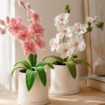 Two crocheted orchid plants — one pink and one white — in white ceramic pots on a light wood shelf with soft natural window light in a minimal Scandinavian interior setting