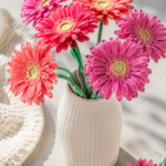 Crocheted gerbera daisies in hot pink coral and magenta yarn arranged in a tall white ribbed ceramic vase on a light grey marble surface with bright natural light and soft shadows