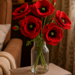 Five crocheted red poppies with black yarn centers in a clear glass vase on stacked hardcover books in a cozy reading nook with warm lamp light in the background