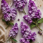 Flat lay of crocheted lilac clusters in deep purple and soft lavender yarn arranged on a weathered white wood surface with vintage linen and dried flowers scattered around