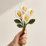A hand holding a small bundle of white crocheted calla lilies with yellow yarn centers and dark green leaves against a clean off-white background in soft side lighting
