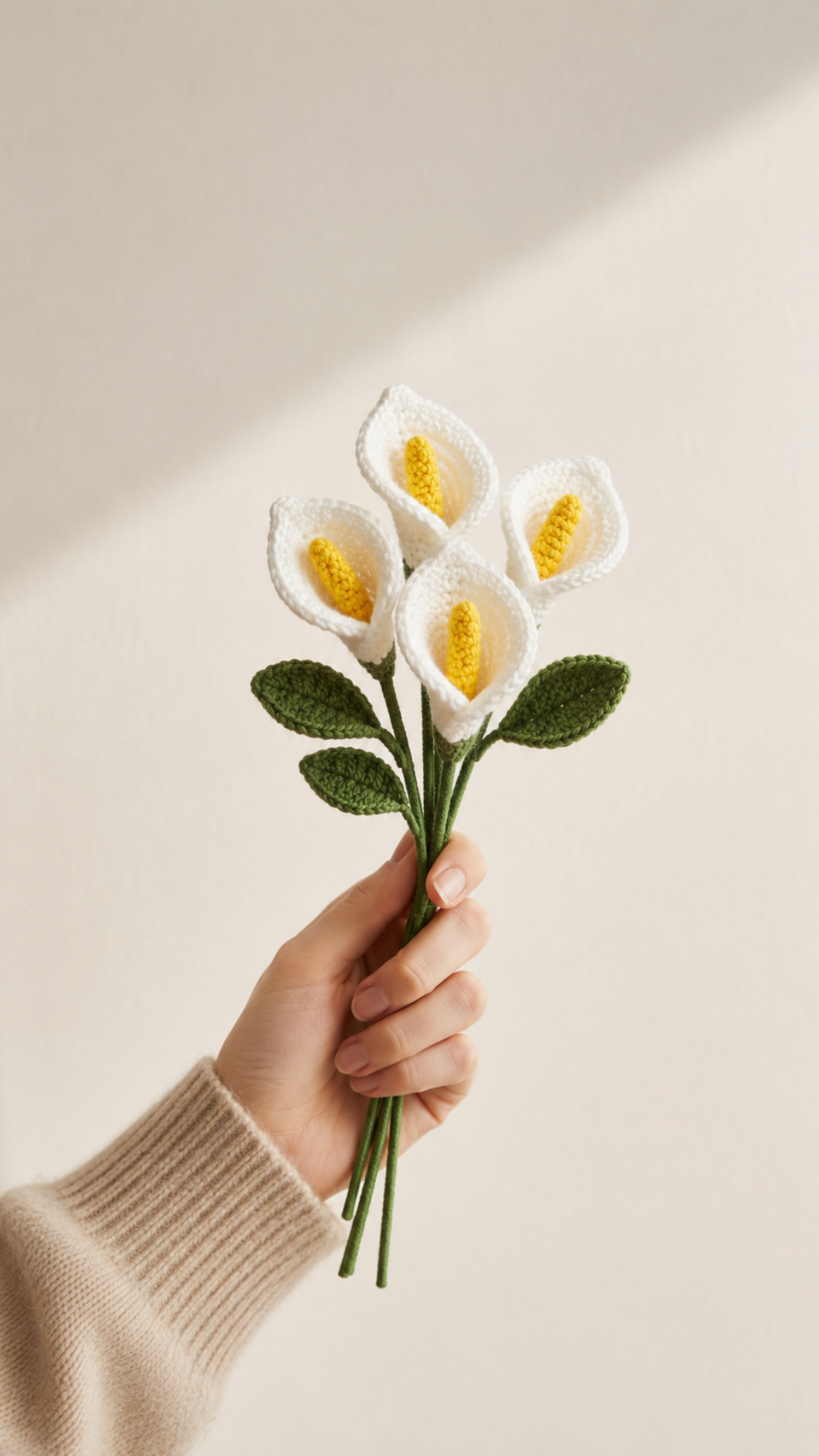 A hand holding a small bundle of white crocheted calla lilies with yellow yarn centers and dark green leaves against a clean off-white background in soft side lighting