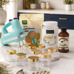 Multiple gold-lidded clear glass jars, an aqua KitchenAid hand mixer, organic brown sugar bag, coconut oil jar, and vanilla extract bottle arranged on white quartz countertops with navy blue cabinetry
