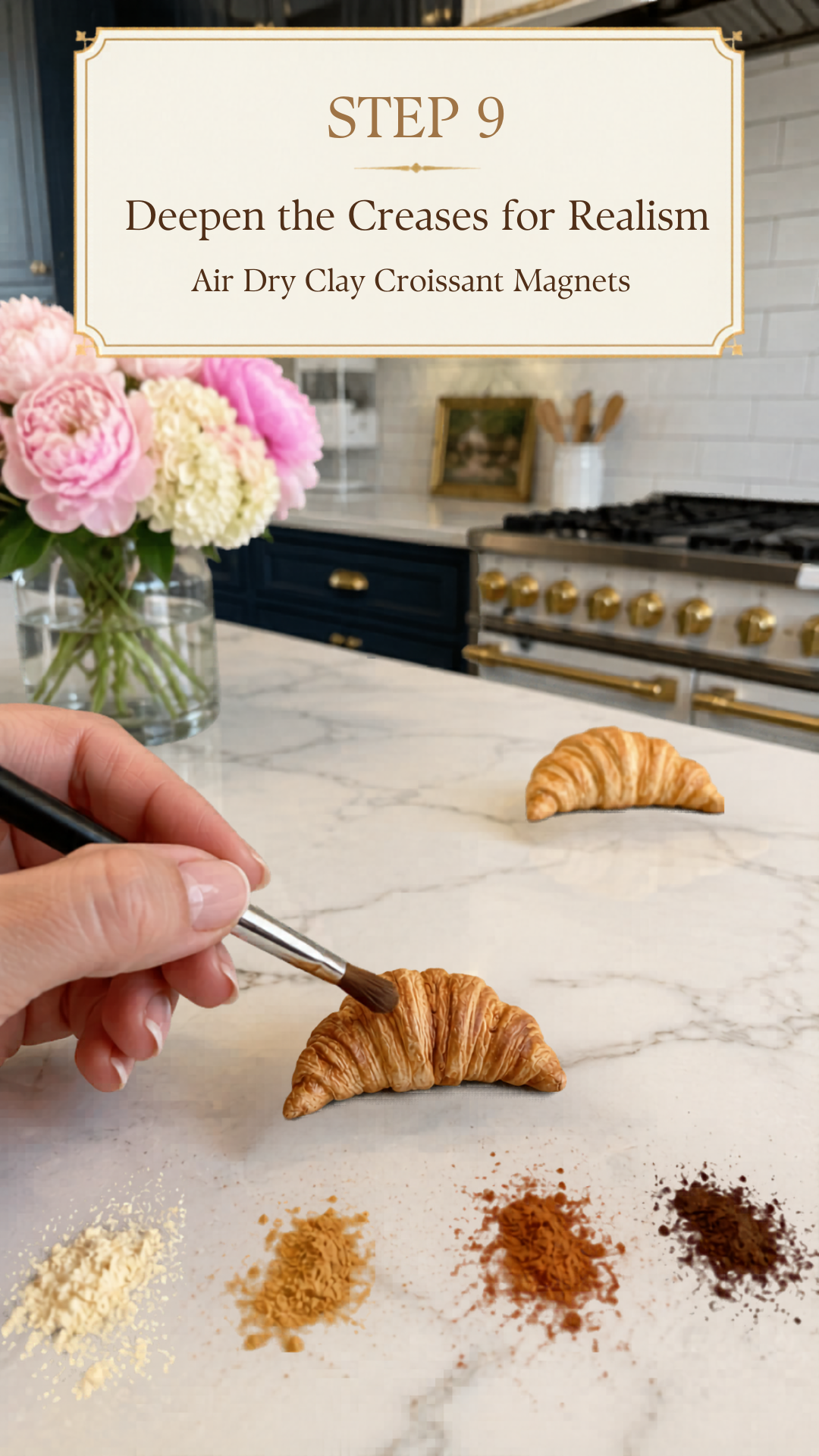 Hand holding a fine black-handled detail brush applying dark chocolate brown dry pastel powder into the layer crevices of a richly golden realistic air dry clay croissant on a white marble countertop, second more lightly colored clay croissant in background, four piles of dry pastel powder in pale cream golden tan rust brown and dark chocolate brown arranged in foreground, pink peonies and white hydrangeas in a glass vase to the left, navy blue kitchen cabinetry with brushed gold hardware and professional gold-knob range visible in background, step nine of air dry clay croissant magnets tutorial deepening creases for realism on jenniferdawn.ca