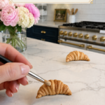 Hand holding a black-handled fine detail brush applying pale cream dry pastel powder highlights to the raised top ridges of a richly golden realistic air dry clay croissant on a white marble countertop, second fully colored clay croissant resting in the background, three piles of dry pastel powder in pale cream golden tan and rust brown arranged in the foreground, pink peonies and cream hydrangeas in a clear glass vase to the upper left, navy blue kitchen cabinetry with brushed gold hardware and professional white range with gold knobs visible in background, step ten of air dry clay croissant magnets tutorial adding pale highlight pastel to raised ridges for dimension on jenniferdawn.ca