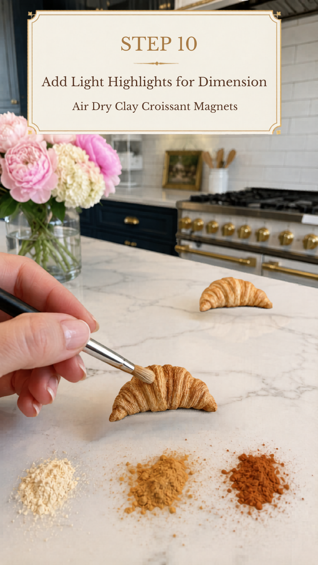 Hand holding a black-handled fine detail brush applying pale cream dry pastel powder highlights to the raised top ridges of a richly golden realistic air dry clay croissant on a white marble countertop, second fully colored clay croissant resting in the background, three piles of dry pastel powder in pale cream golden tan and rust brown arranged in the foreground, pink peonies and cream hydrangeas in a clear glass vase to the upper left, navy blue kitchen cabinetry with brushed gold hardware and professional white range with gold knobs visible in background, step ten of air dry clay croissant magnets tutorial adding pale highlight pastel to raised ridges for dimension on jenniferdawn.ca