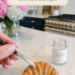 Hand holding a black-handled flat brush applying clear matte varnish to a richly golden realistic air dry clay croissant on a white marble countertop, open glass jar labeled clear varnish matte beside the croissant, pink peonies and blue and white hydrangeas in a clear glass vase to the upper left, navy blue kitchen cabinetry with brushed gold hardware and professional white range with gold knobs visible in background, step eleven of air dry clay croissant magnets tutorial sealing dry pastel color with clear matte varnish on jenniferdawn.ca