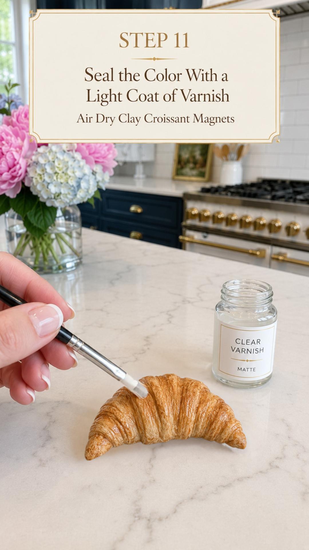 Hand holding a black-handled flat brush applying clear matte varnish to a richly golden realistic air dry clay croissant on a white marble countertop, open glass jar labeled clear varnish matte beside the croissant, pink peonies and blue and white hydrangeas in a clear glass vase to the upper left, navy blue kitchen cabinetry with brushed gold hardware and professional white range with gold knobs visible in background, step eleven of air dry clay croissant magnets tutorial sealing dry pastel color with clear matte varnish on jenniferdawn.ca