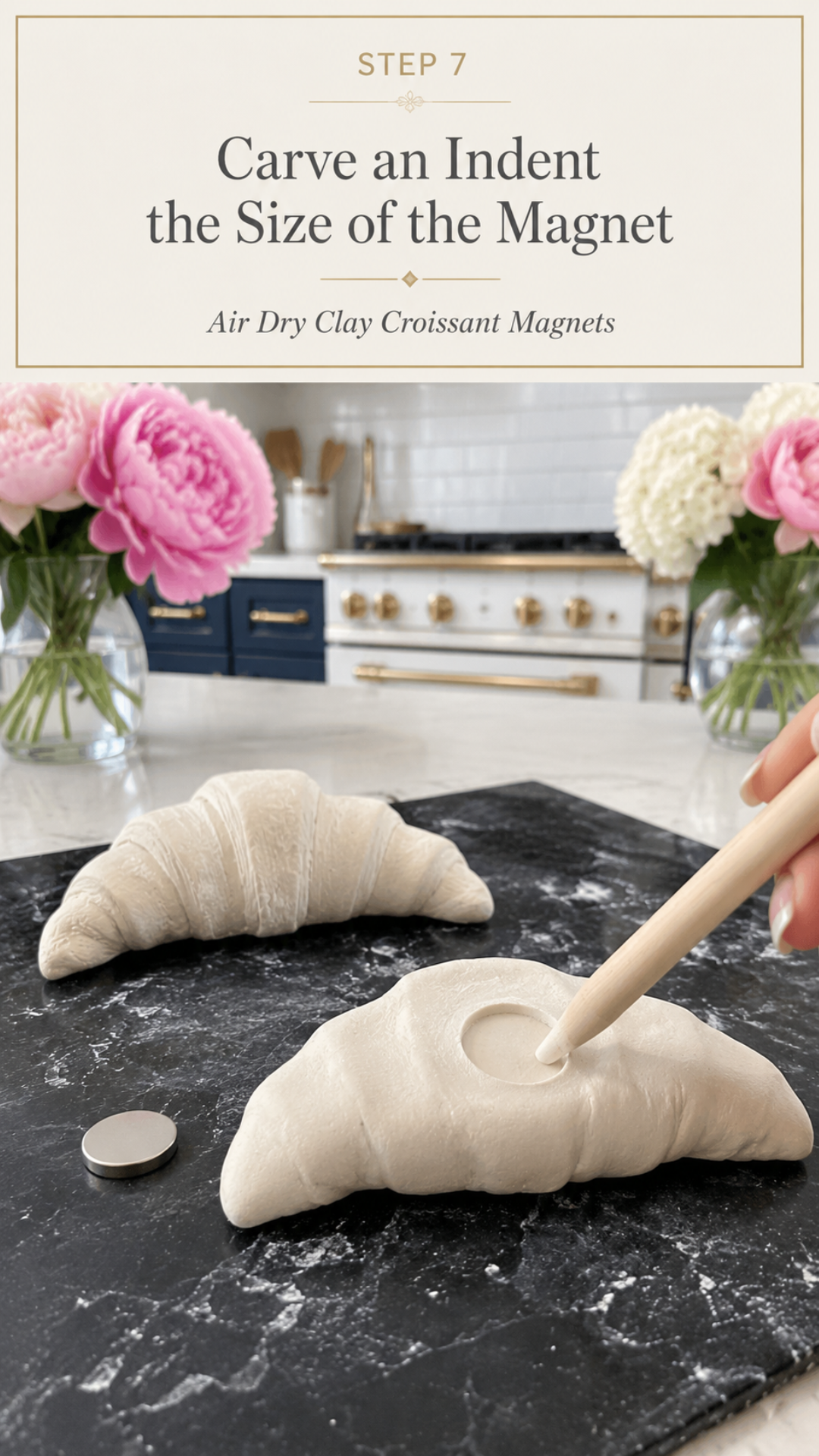 Close view of a hand using a pointed wooden dowel to press a circular indent into the flat back of a large raw white air dry clay croissant resting on a dark slate board, small round silver disc magnet resting on the slate beside the croissant, second unshaped raw clay croissant in background on white quartz countertop, pink peonies in glass vases flanking the scene, navy blue kitchen cabinetry with brushed gold hardware and white professional range with gold knobs and subway tile backsplash, step seven alternate view of air dry clay croissant magnets tutorial carving magnet cavity on jenniferdawn.ca