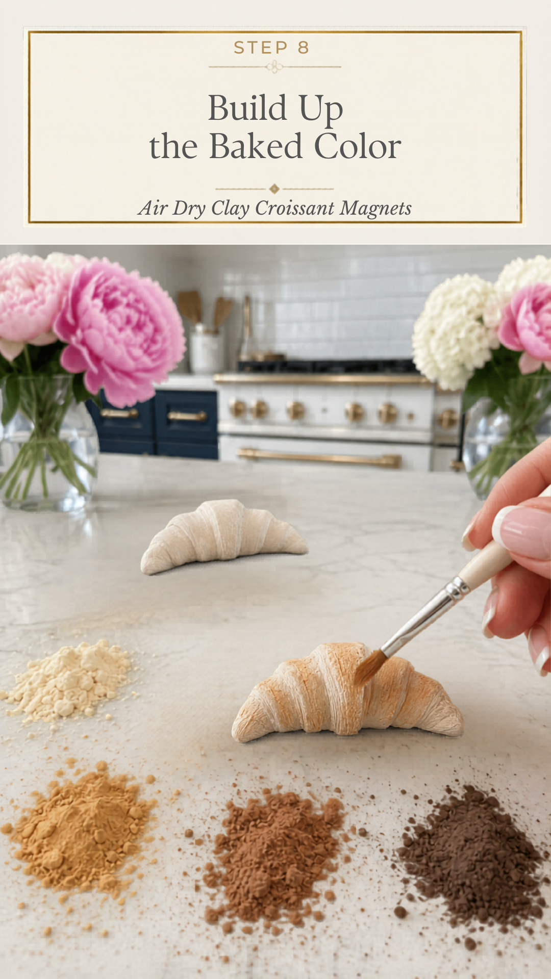 Hand using a flat paintbrush to apply warm golden tan dry pastel powder to a white air dry clay croissant on a white quartz countertop, second uncolored white clay croissant in background, four piles of dry pastel powder in cream golden tan medium brown and dark chocolate brown arranged in foreground, pink peonies and white hydrangeas in glass vases flanking the scene, navy blue kitchen cabinetry with brushed gold hardware and professional gold-knob range with white subway tile backsplash visible, step eight of air dry clay croissant magnets tutorial building up baked color with dry pastels on jenniferdawn.ca