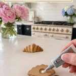 Hand squeezing E6000 industrial strength adhesive from a small grey tube into the circular carved indent on the flat back of a golden colored air dry clay croissant lying face-down on a white quartz countertop, small round silver disc magnet resting beside the croissant ready to be pressed in, second fully finished golden clay croissant standing upright in the background, pink peonies in a glass vase to the left and blue and white hydrangeas in a glass vase to the right, navy blue kitchen cabinetry with brushed gold hardware and professional white range with gold knobs and white subway tile backsplash visible in background, step twelve of air dry clay croissant magnets tutorial gluing the magnet into the indent with E6000 on jenniferdawn.ca