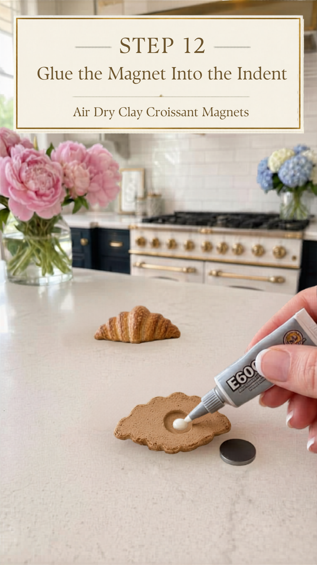 Hand squeezing E6000 industrial strength adhesive from a small grey tube into the circular carved indent on the flat back of a golden colored air dry clay croissant lying face-down on a white quartz countertop, small round silver disc magnet resting beside the croissant ready to be pressed in, second fully finished golden clay croissant standing upright in the background, pink peonies in a glass vase to the left and blue and white hydrangeas in a glass vase to the right, navy blue kitchen cabinetry with brushed gold hardware and professional white range with gold knobs and white subway tile backsplash visible in background, step twelve of air dry clay croissant magnets tutorial gluing the magnet into the indent with E6000 on jenniferdawn.ca