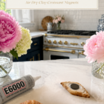 Two realistic golden air dry clay croissants on a white quartz countertop beside a tube of E6000 industrial strength clear self-leveling waterproof adhesive, one croissant lying flat showing a round dark disc magnet freshly glued into the carved back indent, second fully finished croissant standing upright beside it, crystal cut glass vases with pink peonies and cream hydrangeas flanking the scene on both sides, navy blue kitchen cabinetry with brushed gold hardware and professional white range with gold knobs and white subway tile backsplash visible in background, step thirteen of air dry clay croissant magnets tutorial letting E6000 adhesive cure completely before use on jenniferdawn.ca