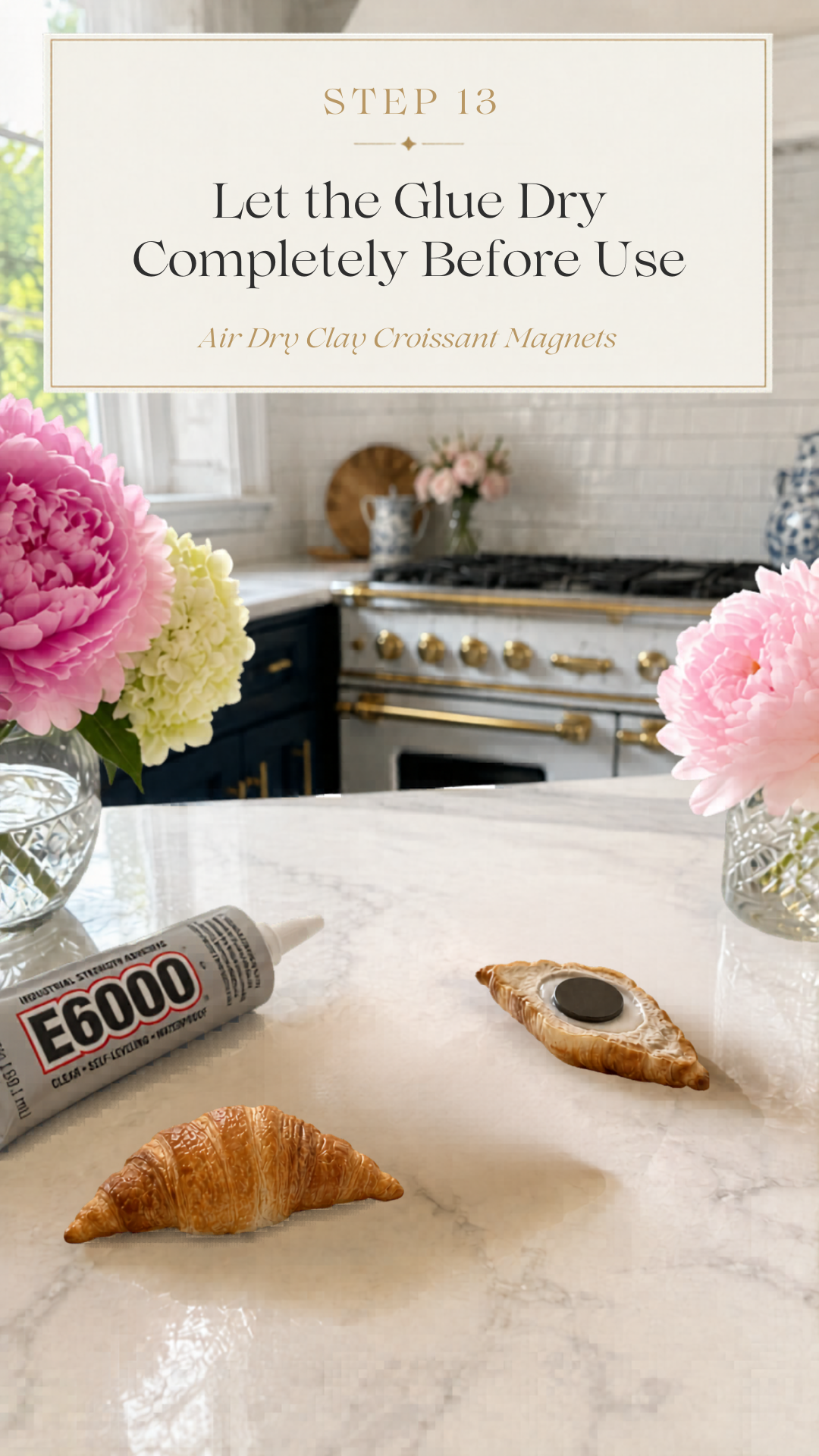 Two realistic golden air dry clay croissants on a white quartz countertop beside a tube of E6000 industrial strength clear self-leveling waterproof adhesive, one croissant lying flat showing a round dark disc magnet freshly glued into the carved back indent, second fully finished croissant standing upright beside it, crystal cut glass vases with pink peonies and cream hydrangeas flanking the scene on both sides, navy blue kitchen cabinetry with brushed gold hardware and professional white range with gold knobs and white subway tile backsplash visible in background, step thirteen of air dry clay croissant magnets tutorial letting E6000 adhesive cure completely before use on jenniferdawn.ca