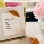 Three finished realistic golden air dry clay croissant magnets displayed on a white marble countertop, one croissant magnet holding a handwritten market list reading eggs butter honey peonies on a torn paper note clipped to a white gold-framed magnetic board, two additional finished croissants resting on the countertop beside it, crystal cut glass vase with pink peonies to the right, navy blue kitchen cabinetry with brushed gold hardware visible in background, completed step fourteen of air dry clay croissant magnets tutorial final result display on jenniferdawn.ca