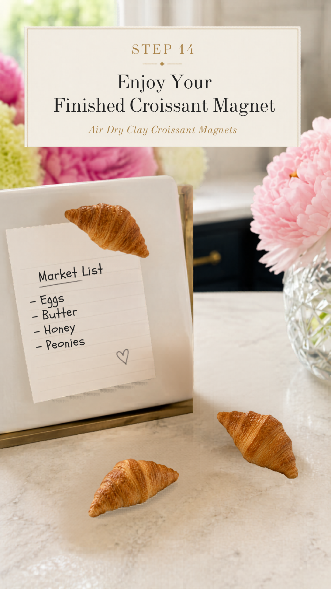 Three finished realistic golden air dry clay croissant magnets displayed on a white marble countertop, one croissant magnet holding a handwritten market list reading eggs butter honey peonies on a torn paper note clipped to a white gold-framed magnetic board, two additional finished croissants resting on the countertop beside it, crystal cut glass vase with pink peonies to the right, navy blue kitchen cabinetry with brushed gold hardware visible in background, completed step fourteen of air dry clay croissant magnets tutorial final result display on jenniferdawn.ca