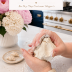 Two hands kneading a large piece of white air dry clay on a white quartz countertop dusted with cornstarch, clay sculpting tools and a small finished raw clay croissant on a ceramic plate beside the clay, white ribbed vase with pink peonies and white hydrangeas in background, navy blue kitchen cabinetry with brushed gold hardware and professional gold-knob range visible, step one of air dry clay croissant magnets tutorial conditioning the clay on jenniferdawn.ca