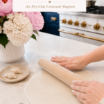 Two hands using a wooden rolling pin to roll out a large round sheet of white air dry clay on a white quartz countertop dusted with cornstarch, clay sculpting tools beside the slab, small finished raw clay croissant on a ceramic plate to the left, white ribbed vase with pink peonies and white hydrangeas in background, navy blue kitchen cabinetry with brushed gold hardware and professional gold-knob range visible, step two of air dry clay croissant magnets tutorial rolling the clay evenly on jenniferdawn.ca