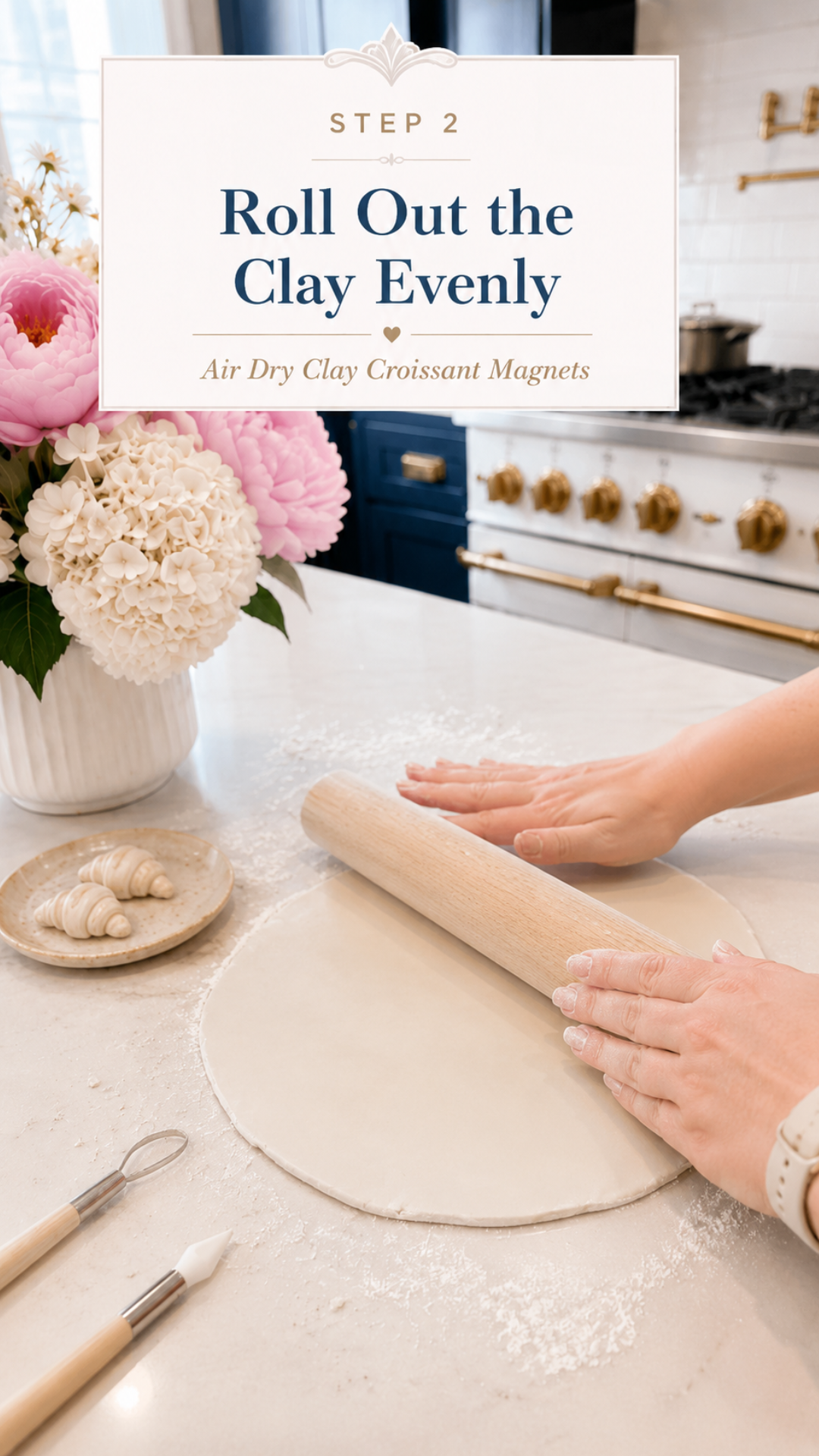 Two hands using a wooden rolling pin to roll out a large round sheet of white air dry clay on a white quartz countertop dusted with cornstarch, clay sculpting tools beside the slab, small finished raw clay croissant on a ceramic plate to the left, white ribbed vase with pink peonies and white hydrangeas in background, navy blue kitchen cabinetry with brushed gold hardware and professional gold-knob range visible, step two of air dry clay croissant magnets tutorial rolling the clay evenly on jenniferdawn.ca