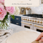 Hand placing a kraft cardboard triangle template onto a large round rolled sheet of white air dry clay on a white marble countertop, clay sculpting tools and a linen napkin beside the slab, pink peonies and white hydrangeas in a clear glass vase to the left, navy blue kitchen cabinetry with brushed gold hardware and professional gold-knob range with white subway tile backsplash visible in background, step three of air dry clay croissant magnets tutorial using a template to cut triangles on jenniferdawn.ca