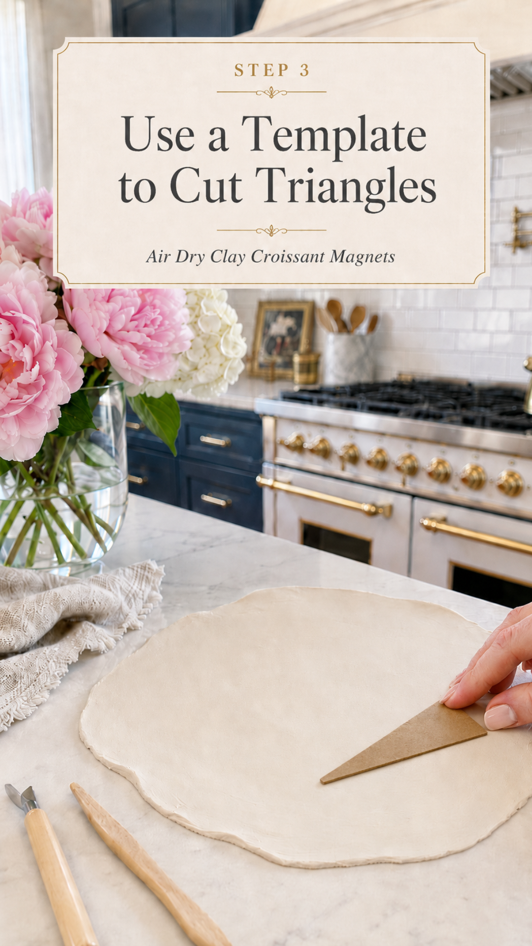 Hand placing a kraft cardboard triangle template onto a large round rolled sheet of white air dry clay on a white marble countertop, clay sculpting tools and a linen napkin beside the slab, pink peonies and white hydrangeas in a clear glass vase to the left, navy blue kitchen cabinetry with brushed gold hardware and professional gold-knob range with white subway tile backsplash visible in background, step three of air dry clay croissant magnets tutorial using a template to cut triangles on jenniferdawn.ca