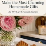 Three finished realistic air dry clay croissant magnets displayed on a white marble countertop beside a small white framed board with a handwritten market list, pink peonies and hydrangeas in a crystal vase, navy blue kitchen cabinetry with gold hardware in background, completed air dry clay croissant magnets project styled as a handmade gift, jenniferdawn.ca
