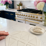 Overhead view of a large round white air dry clay slab cut into ten triangles in a pinwheel pattern on a white quartz countertop, hand resting on the edge of the slab, cardboard triangle template and clay knife beside the slab, two raw rolled clay croissants on a gold-rimmed ceramic plate to the right, pink peonies and white and blue hydrangeas in glass vases, navy blue kitchen cabinetry with brushed gold hardware and professional gold-knob range with white subway tile backsplash in background, step four of air dry clay croissant magnets tutorial cutting multiple triangles from one slab on jenniferdawn.ca