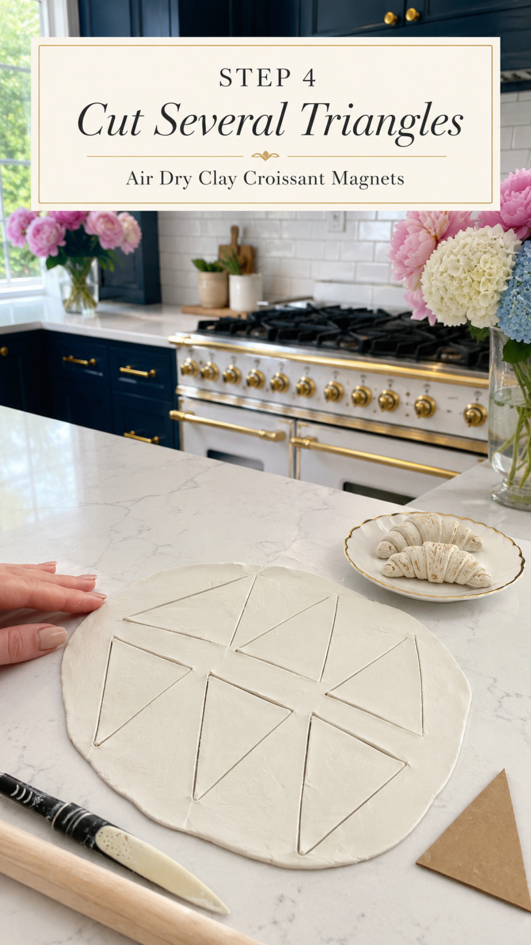 Overhead view of a large round white air dry clay slab cut into ten triangles in a pinwheel pattern on a white quartz countertop, hand resting on the edge of the slab, cardboard triangle template and clay knife beside the slab, two raw rolled clay croissants on a gold-rimmed ceramic plate to the right, pink peonies and white and blue hydrangeas in glass vases, navy blue kitchen cabinetry with brushed gold hardware and professional gold-knob range with white subway tile backsplash in background, step four of air dry clay croissant magnets tutorial cutting multiple triangles from one slab on jenniferdawn.ca