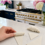 Hand rolling a white air dry clay triangle into a croissant shape on a white quartz countertop, three additional unrolled clay triangles and one fully rolled raw clay croissant visible nearby, clay sculpting tool and wooden rolling pin in foreground, pink and white flower arrangements in glass vases in background, navy blue kitchen cabinetry with brushed gold hardware and professional gold-knob range with white subway tile backsplash visible, step five of air dry clay croissant magnets tutorial rolling triangles into croissant shapes on jenniferdawn.ca