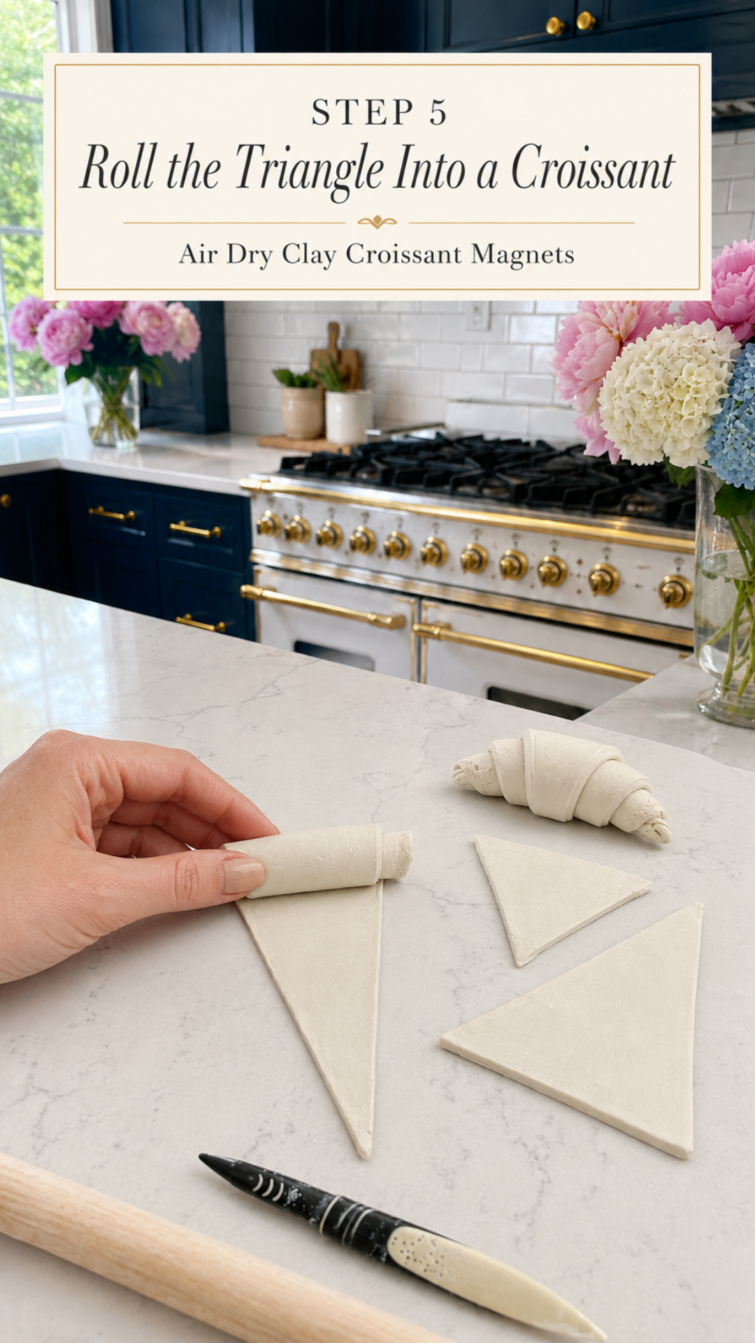 Hand rolling a white air dry clay triangle into a croissant shape on a white quartz countertop, three additional unrolled clay triangles and one fully rolled raw clay croissant visible nearby, clay sculpting tool and wooden rolling pin in foreground, pink and white flower arrangements in glass vases in background, navy blue kitchen cabinetry with brushed gold hardware and professional gold-knob range with white subway tile backsplash visible, step five of air dry clay croissant magnets tutorial rolling triangles into croissant shapes on jenniferdawn.ca
